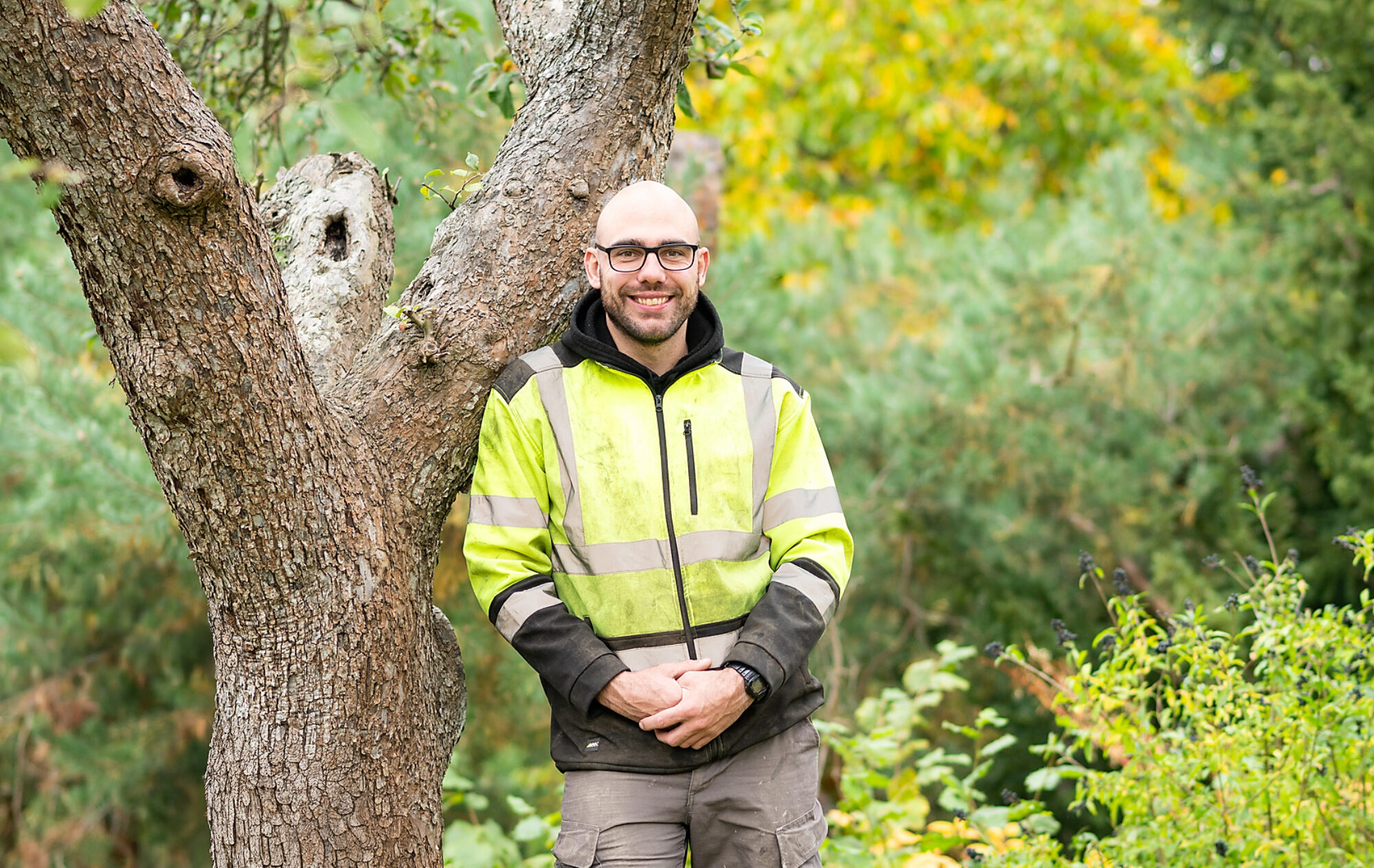Auf dem Bild sieht man David Böhm, der sich in Arbeitskleidung entspannt an einen alten Baum mit grober Rinde lehnt und in die Kamera lächelt. Im Hintergrund sieht man ganz verschwommen einen belaubten, grünen Garten zu beginn des Herbstes.